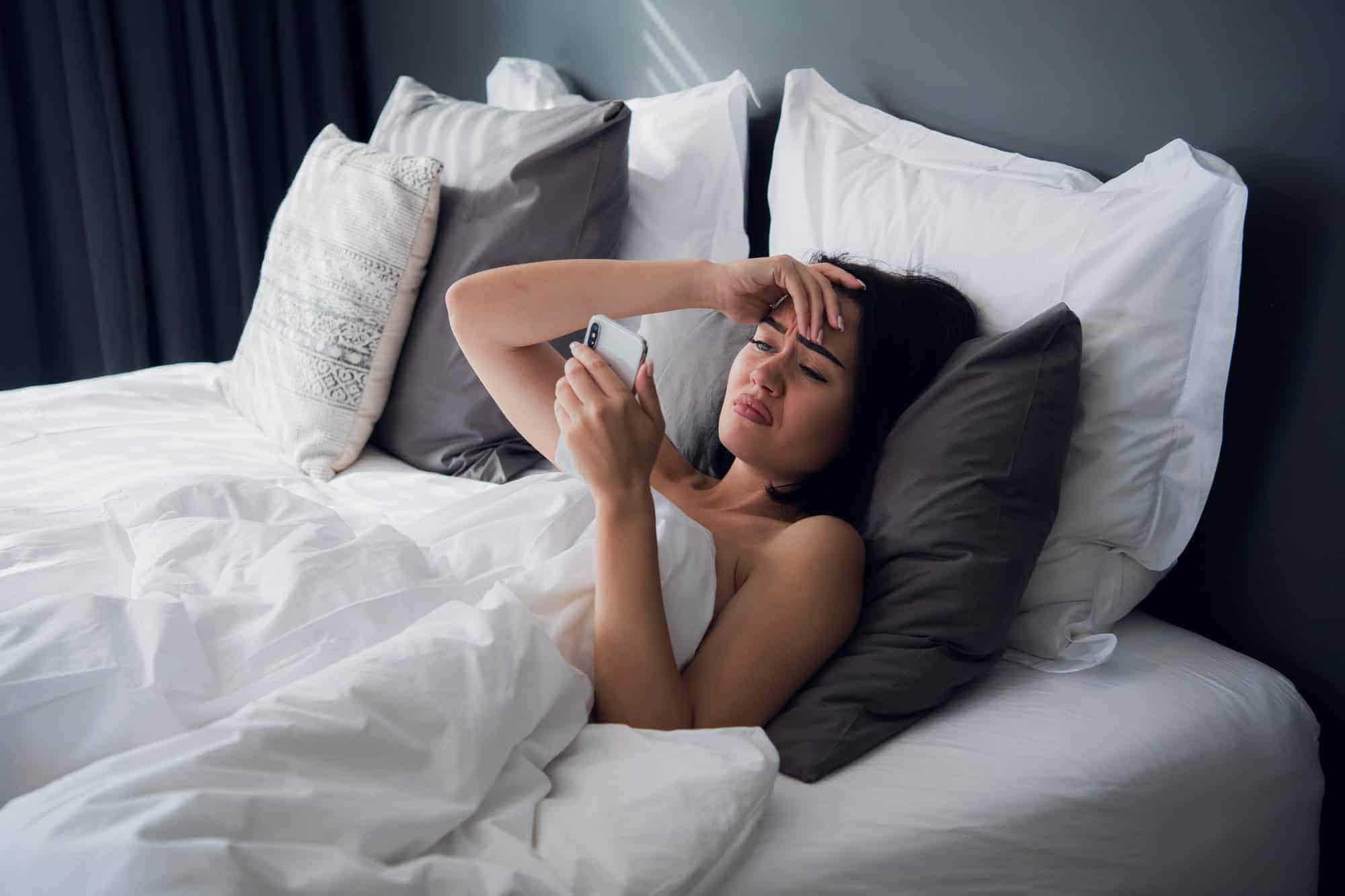 Image of frustrated upset woman 20s with dark hair lying in bed on white pillow after sleep and